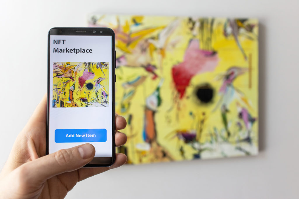 A hand holding a phone displaying an artwork on its screen, with a physical artwork hanging on the wall in the background. NFT marketplace interface is visible at the top, offering the option to buy the artwork.